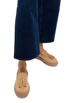 Voeten met beige suède loafers met kwastjes, gecombineerd met donkere wijde denim jeans, tegen een witte achtergrond.