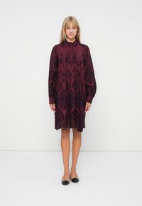 Burgundy dress with black embroidery, long sleeves, pleated detail at neckline, and knee-length hem. Worn with black flat shoes.