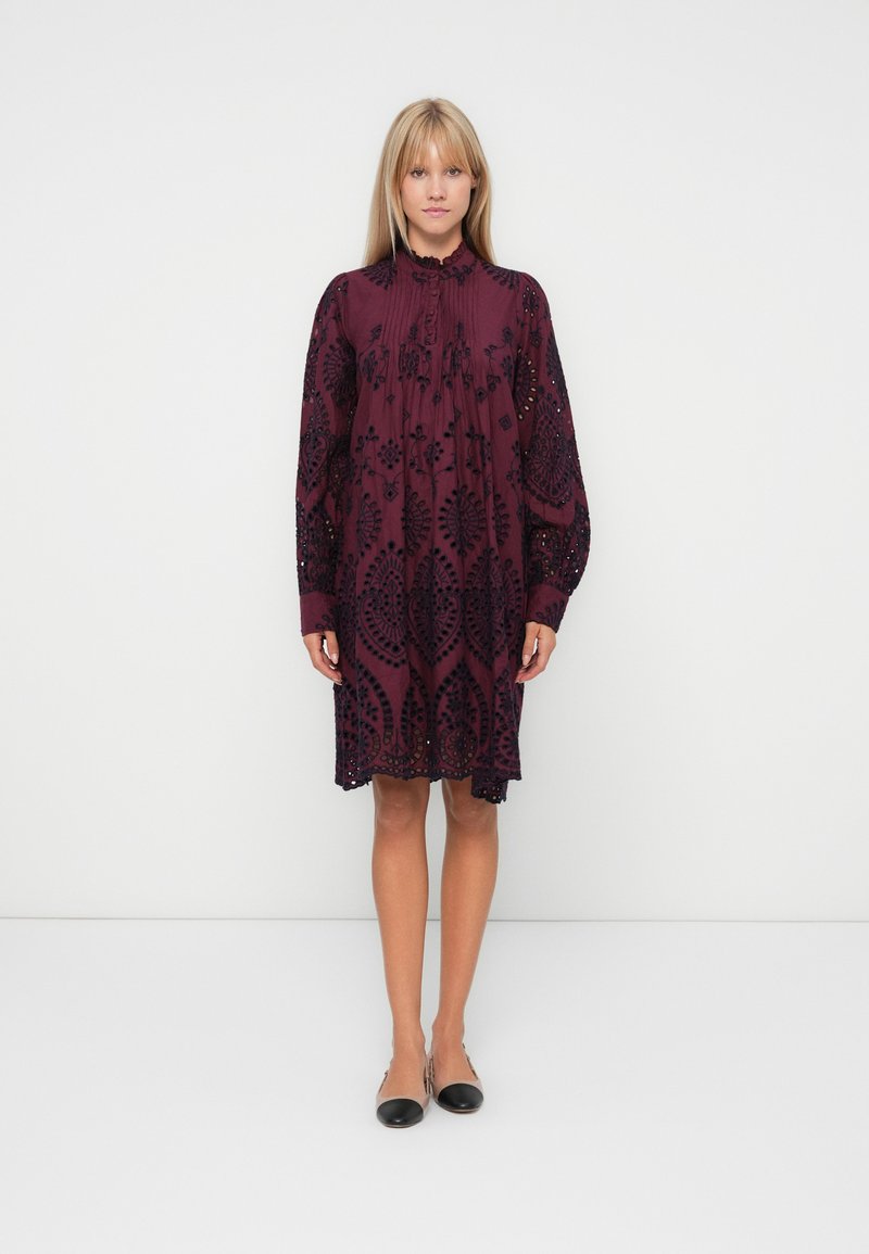 Burgundy dress with black embroidery, long sleeves, pleated detail at neckline, and knee-length hem. Worn with black flat shoes.