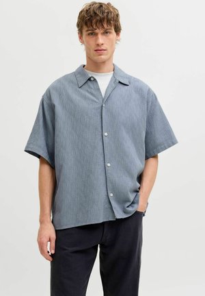 Młody mężczyzna z potarganymi włosami, ubrany w jasnoniebieską koszulę z krótkim rękawem zapinaną na guziki, na białej koszulce i ciemnych spodniach, na jednolitym tle.