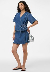 Denim wrap-toppi lyhyillä hihoilla ja nyöriyksityiskohdalla, yhdistettynä yhteensopivaan denimihameseen. Hopeinen käsilaukku, mustat flip-flopit.