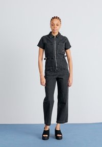 Svart denim jumpsuit med korte ermer, glidelås foran, to brystlommer og justerbar midje. Sammen med svarte platåsko.