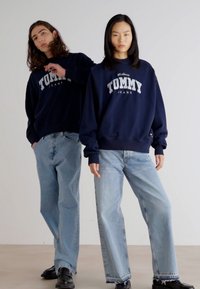 Sudaderas azul marino con "TOMMY JEANS" impresas en el frente, combinadas con jeans de pierna ancha azul claro y zapatos negros.