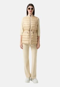 Beige Steppweste mit hohem Kragen, Kordelzug in der Taille und zwei Reißverschlusstaschen, kombiniert mit cremfarbenen Hosen und weißen Sneakern.