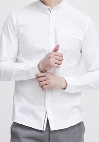 Hvid skjorte med knaplukning og båndkrave, lange ærmer og lomme på brystet. Stof ser glat og skarpt ud, fremhævet af detaljeret syning.