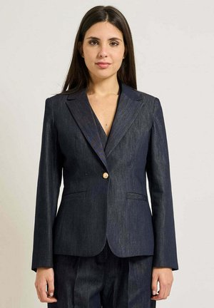 Donna con lunghi capelli scuri che indossa un blazer di jeans scuro con un solo bottone dorato, in piedi davanti a uno sfondo chiaro e semplice.