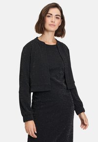 Giacca nera corta con maniche lunghe, realizzata in tessuto testurizzato con fili metallici, abbinata a un vestito nero coordinato.