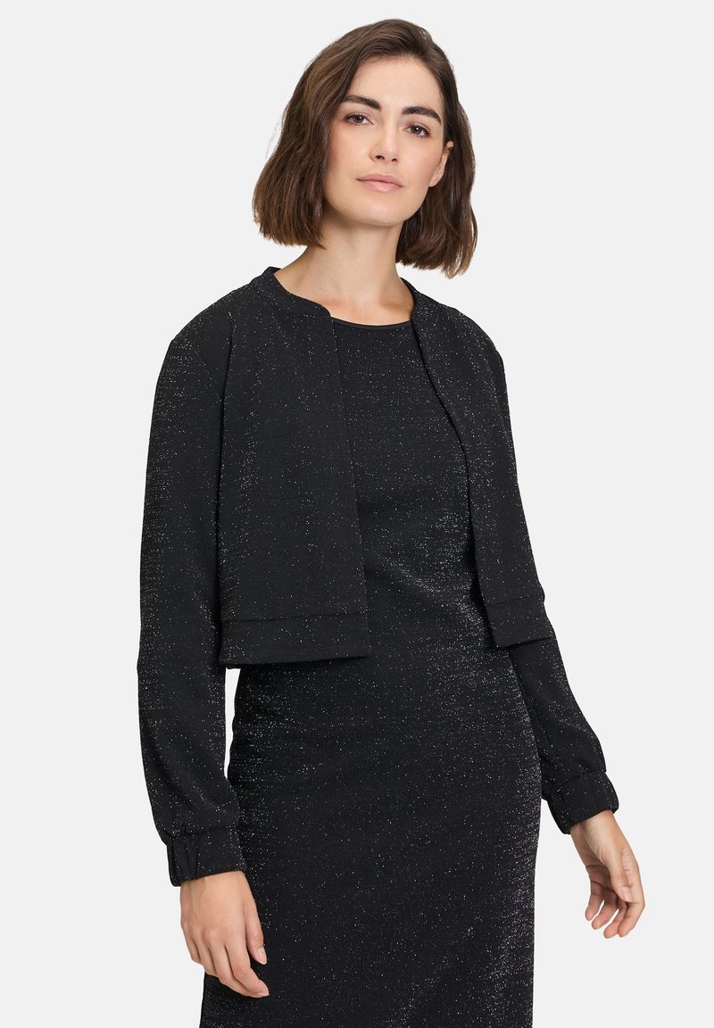 Giacca nera corta con maniche lunghe, realizzata in tessuto testurizzato con fili metallici, abbinata a un vestito nero coordinato.