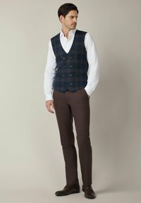 Granatowa kamizelka w kratę z podwójnymi guzikami, biała koszula pod spodem, brązowe spodnie i ciemne buty, prezentująca dopasowany fason i kontrastujące wzory.