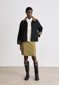 Casaco preto com colarinho castanho, detalhes verdes, fecho de zipper e bolsos. Usado sobre um top branco de malha canelada e uma saia lápis verde, combinado com botas pretas.