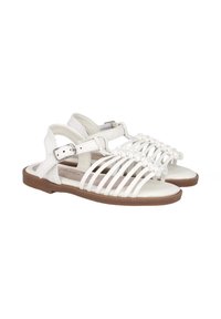 Sandalias blancas con tiras y suela plana marrón. Presentan correas trenzadas en la parte delantera y un cierre de hebilla en el lateral. Material de cuero suave.