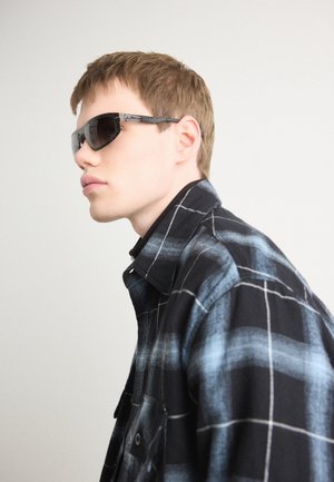 Camisa de cuadros en negro y azul con cuello abotonado, llevada con gafas de sol oscuras y de gran tamaño, que presentan un diseño elegante y angular.