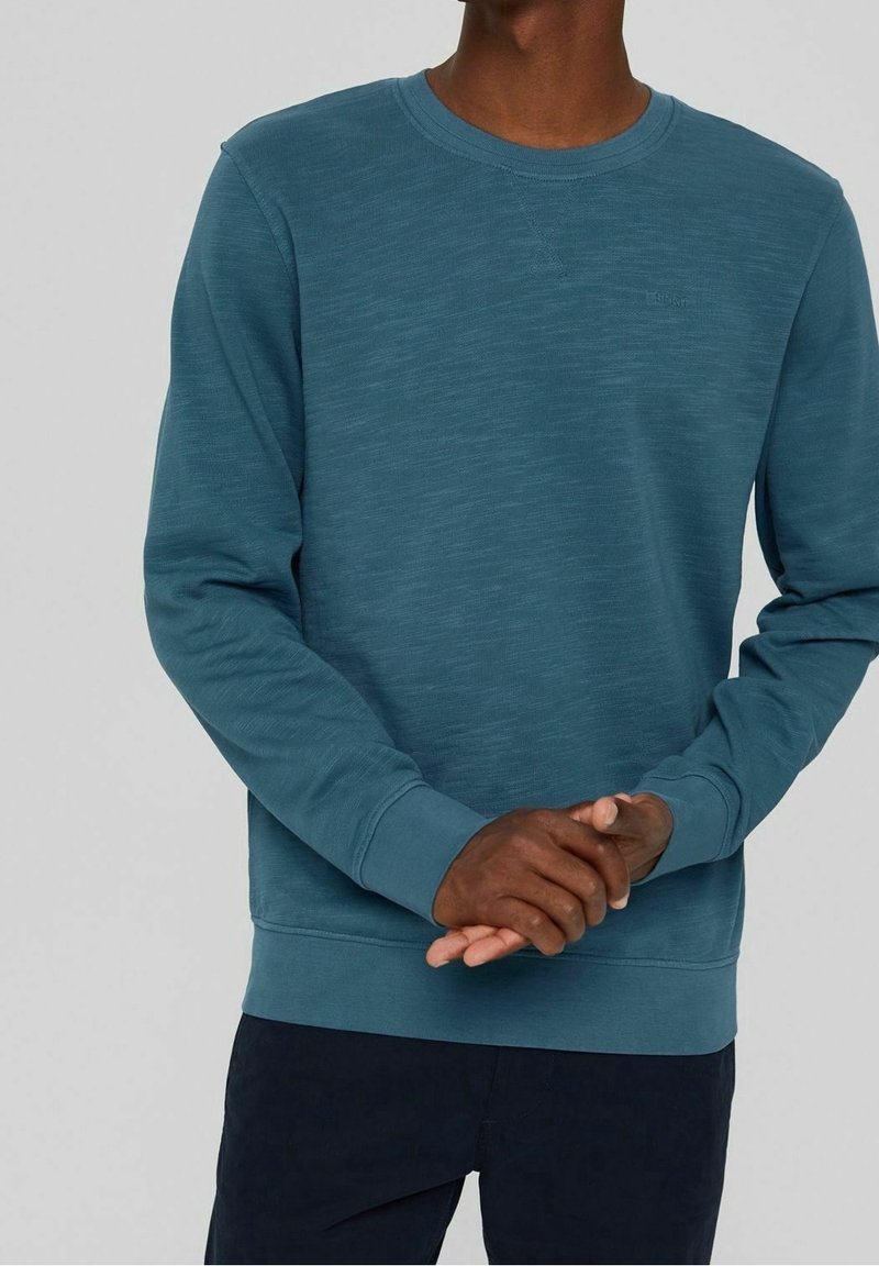 Sudadera de cuello redondo en color verde azulado, hecha de tejido texturizado, con puños y dobladillo acanalados; presenta un sutil logotipo en el pecho.