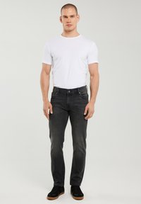 Egy férfi áll előre nézve, egyszerű fehér pólót, kifakult fekete farmert és barna talpú fekete tornacipőt visel, világos háttér előtt.