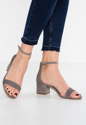 Pies con sandalias de tacón ancho de ante gris con tiras en el tobillo, combinadas con vaqueros azul oscuro con bajos deshilachados sobre un suelo blanco.