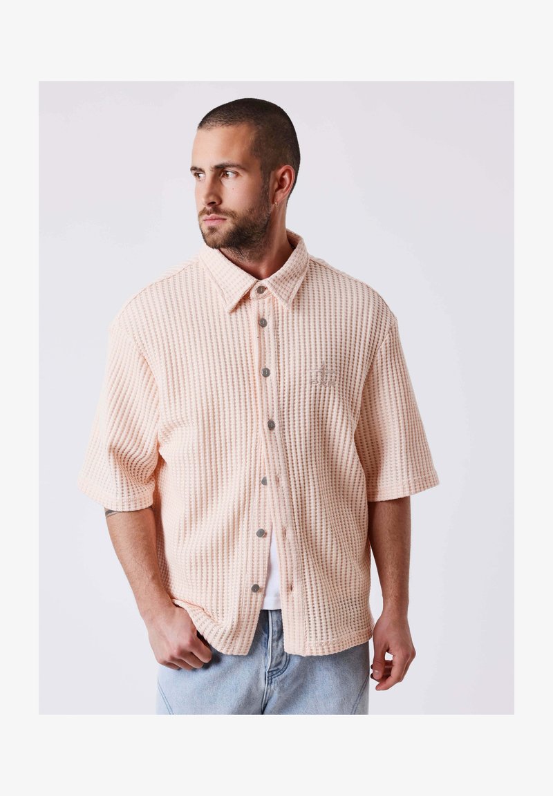 Homme aux cheveux courts et barbe portant une chemise beige en maille gaufrée à manches courtes boutonnée et un jean bleu clair, regardant vers la gauche.