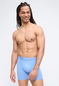 Χαμογελαστός άντρας με μακριά μαλλιά, χωρίς πουκάμισο, φορώντας ανοιχτό μπλε boxer briefs Calvin Klein, στέκεται μπροστά σε απλό φωτεινό φόντο.