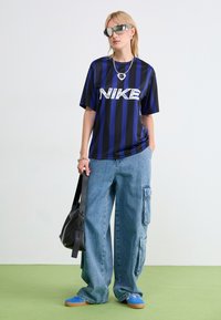 Camiseta Nike a rayas negras y azules, de corte holgado, combinada con pantalones cargo de denim anchos. Los accesorios incluyen una cadena de plata y gafas de sol reflectantes.