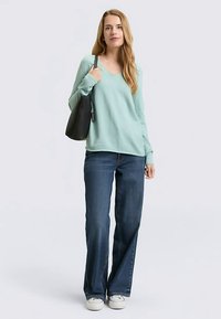 Hellgrüner V-Ausschnitt-Pullover, locker sitzend, kombiniert mit dunkelblauen Jeans mit geradem Bein. Das Model hält eine schwarze Handtasche. Weiße Turnschuhe runden das Outfit ab.