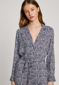 Cvečni kombinezon z mornarsko modro podlago in rožnatimi ter belimi cvetličnimi vzorci, dolgimi rokavi, ovratnikom in vezanim pasom.