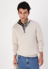 Beige Strickpullover mit einem halben Reißverschluss-Kragen, ausgestattet mit einem schwarzen Reißverschluss und gerippten Bündchen und Saum, über einem karierten Hemd getragen; blaue Jeans.