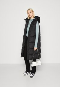 Zwarte puffer gilet met een capuchon, gestructureerd oppervlak, zijzakken, gecombineerd met een lichtblauwe geribde sweater, zwarte broek en zwart-witte sneakers.
