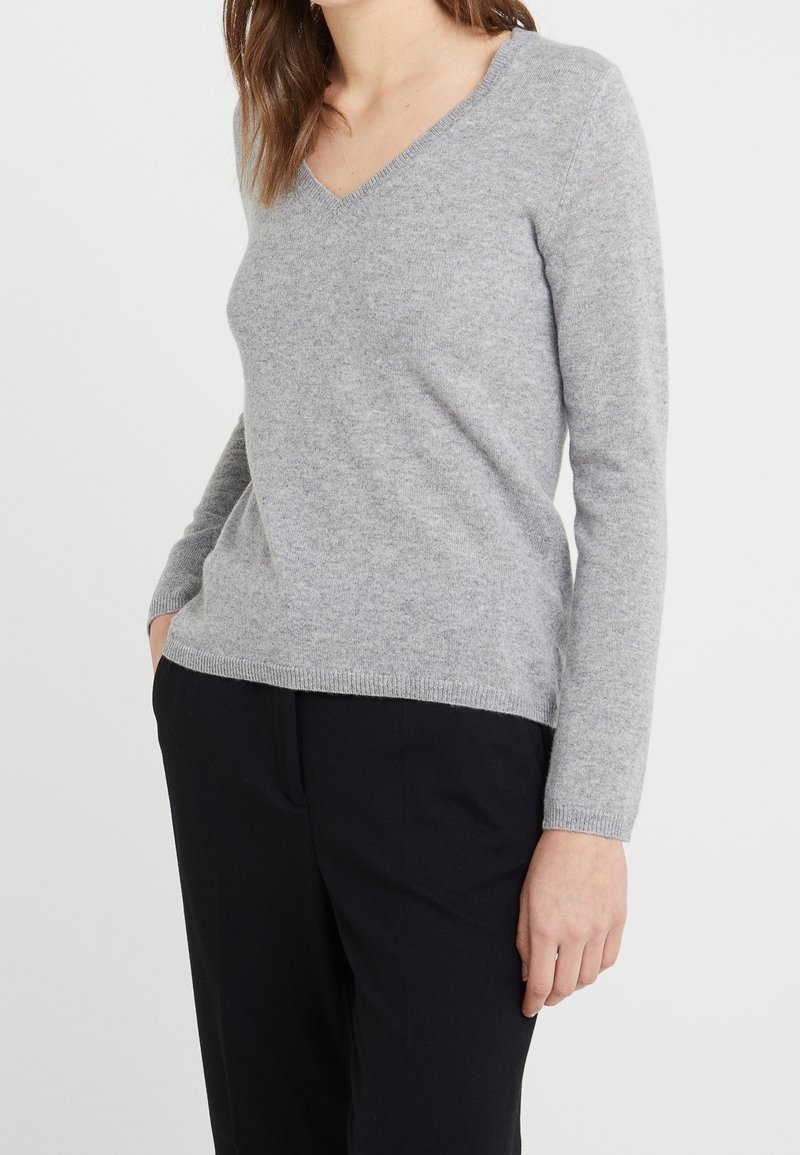 Femme portant un pull gris clair à col en V et un pantalon noir, debout les mains détendues, sur un fond blanc uni.