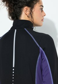 Žena s kudrnatými vlasy v černé a fialové sportovní bundě s vysokým límcem, odrazivým vertikálním pruhem a šitými detaily, směrem vpravo.