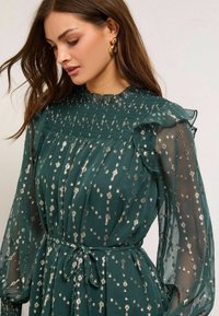 Zelené šaty s priesvitnými dlhými rukávmi, zúženou sukňou, volánovým výstrihom a striebornými kvetinovými detailmi. Uviazaný pás pre definovanie postavy.