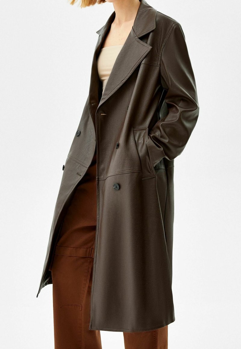 Personne portant un long manteau trench en cuir brun foncé sur un haut bandeau beige et un pantalon large marron, main dans la poche du manteau.