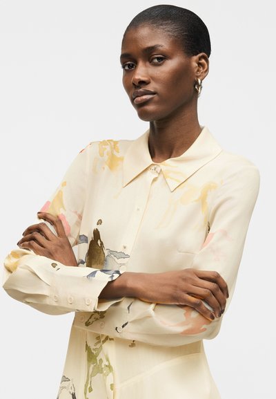 Γυναίκα με σταυρωμένα χέρια, φορώντας κρεμ μπλούζα με υποτονικά πολύχρωμα αφηρημένα σχέδια και χρυσούς κρίκους σκουλαρίκια, μπροστά σε απλό φόντο.