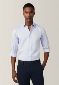 Camisa de botones azul claro con las mangas arremangadas, fabricada con un tejido texturizado, combinada con pantalones navy que tienen un cierre de un solo botón.