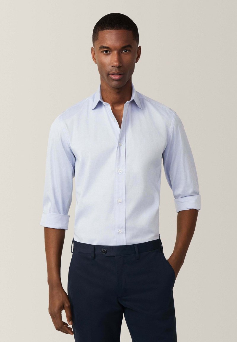 Camisa de botones azul claro con las mangas arremangadas, fabricada con un tejido texturizado, combinada con pantalones navy que tienen un cierre de un solo botón.