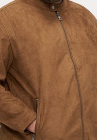 Veste en daim marron avec fermeture éclair à l'avant, poche latérale zippée et détail de col à bouton-pression, portée par une personne avec une main dans la poche.