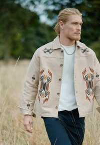 Beige broderad jacka med geometriska mönster i brunt, svart och orange. Knapp-up design, krage, bärs över en vit skjorta.