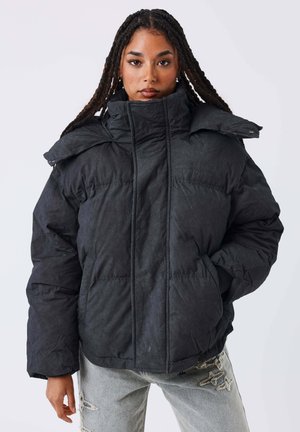 Femme avec de longues tresses portant une doudoune noire et un jean gris clair avec des étoiles brodées, debout sur un fond blanc.