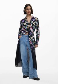 Svart skjorta med blommönster och lång, genomskinlig design. Kombinerad med ljusblå vida jeans. Har en knuten framdel och krage.