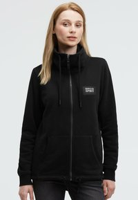 Black zip-up hoodie made of soft fabric, featuring a high collar, drawstrings, and a logo patch on the chest. Large front pockets visible.