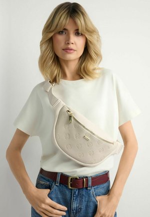 Blondynka w białej koszulce i niebieskich dżinsach z czerwonym paskiem, niosąca beżową torbę na ramię z tłoczonymi logo i złotymi suwakami.