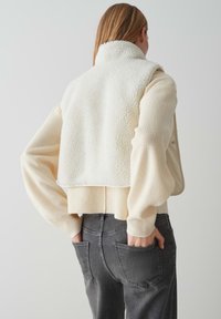 Roomwitte, fluffy fleece vest over een lichtbeige gebreide trui. Grijze jeans met zichtbare achterzakken. Neutrale kleuren en zachte texturen.