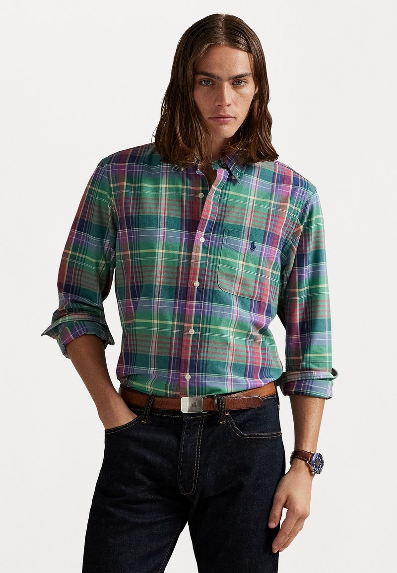 Homme portant une chemise à carreaux multicolore verte et violette avec des manches retroussées, un jean sombre, une ceinture marron et une montre, se tenant devant un fond blanc.