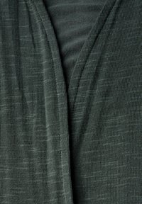 Grön stickad tyg med en subtil textur; har en V-ringning och överlappande paneler för en lagerlook. Lätt och andningsbar.