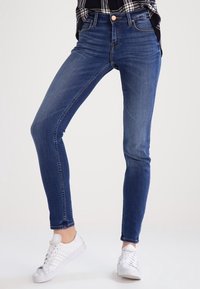 Jeans ajustados de denim azul oscuro, con un talle medio, diseño clásico de cinco bolsillos y costuras sutiles a lo largo de las costuras, combinados con zapatillas blancas.