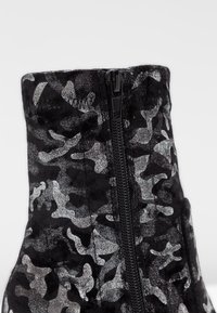 Bottine noire camouflage avec une texture douce, présentant un motif argenté, un col haut et une fermeture éclair noire sur le côté.