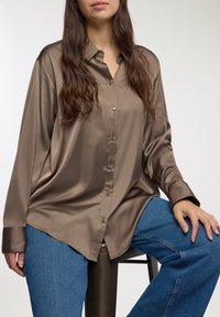Camicia button-up in raso marrone con una vestibilità comoda, maniche lunghe e colletto classico, abbinata a jeans di denim blu.