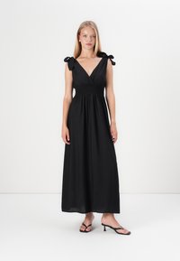 Vestido maxi preto com decote em V, cintura franzida e alças amarradas. Tecido leve. Usado com sandálias de salto pretas.