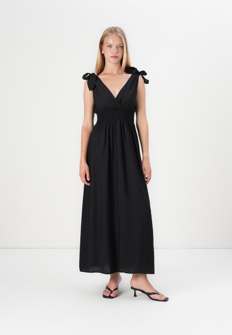 Vestido maxi preto com decote em V, cintura franzida e alças amarradas. Tecido leve. Usado com sandálias de salto pretas.