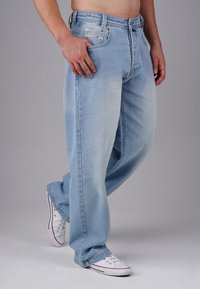 Hellblaue Jeans mit lockerer Passform, ausgestattet mit einem Reißverschluss, Hardware in Kupferfarbe und Details an den Vorderentaschen. Kombiniert mit weißen Sneakern.