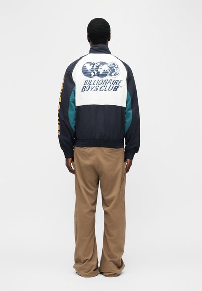 Άνδρας που στέκεται πλάτη φορώντας ένα μπουφάν Billionaire Boys Club σε μπλε ναυτικό και λευκό και μπεζ παντελόνι με φαρδιά πόδια, σε λευκό φόντο.