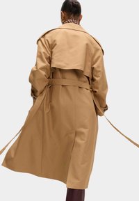 Beige Trenchcoat mit tailliertem Gürtel, doppelter Knopfleiste und Rückenschlitzdetails. Glatte Textur und knielanger Schnitt mit seitlichen Schlitzen.
